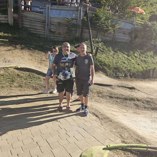 Drei Personen stehen auf einem Lehmweg, einer hält eine Trophäe. Eine Frau steht hinter ihnen und ein Holzzaun im Hintergrund.