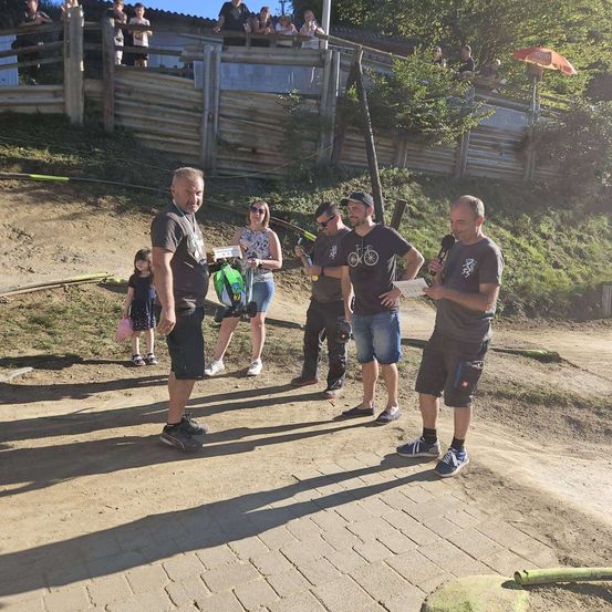 Eine Gruppe von Menschen steht auf einem Erdweg, einer hält ein Mikrofon und spricht. Eine Frau hält eine Kamera und ein Mann hält ein grünes Objekt. Dahinter ist ein Holzzaun und ein Baum zu sehen.