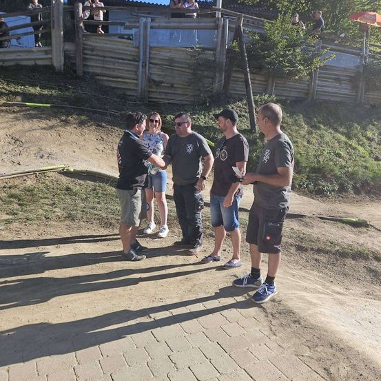 Fünf Personen stehen auf einem Erdweg, zwei Frauen stehen hinter ihnen. Sie tragen alle Sonnenbrillen und Turnschuhe. Hinter ihnen steht ein Holzzaun, auf dem einige Personen stehen.