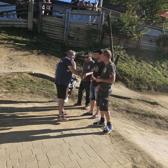 Fünf Männer stehen auf einem staubigen Pfad, zwei halten Getränke, einer hält eine Kamera, mit einem Holzzaun und Zuschauern im Hintergrund.