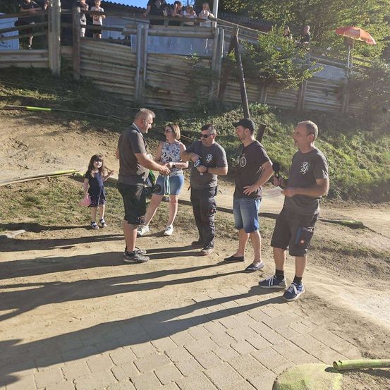 Eine Gruppe von Menschen steht auf einem staubigen Pfad, einer hält eine Kamera. Dahinter trennt ein Holzzaun den Weg von einem Grasbereich mit einem Gebäude. Ein kleines Kind ist ebenfalls anwesend.