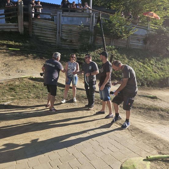 Eine Gruppe von Menschen steht auf einem Lehmweg, mit einem Holzzaun und einem Hügel im Hintergrund. Einige tragen Sonnenbrillen und Turnschuhe.