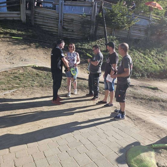 Fünf Personen stehen auf einem Erdweg mit einem Holzzaun und Grünpflanzen im Hintergrund. Ein Mann hält ein Mikrofon und Papier, während ein anderer ein gelbes Objekt in der Hand hat.