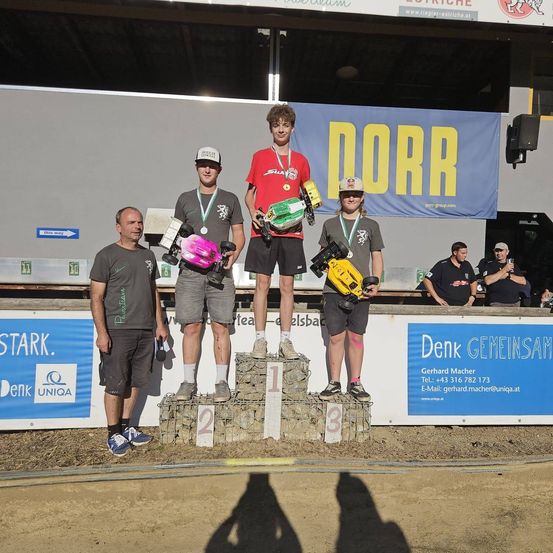 Drei junge Männer stehen auf einem Podium mit Medaillen um den Hals. Sie halten ferngesteuerte Autos. Ein Mann steht links und ein Banner mit dem Wort Dorr ist hinter ihnen.