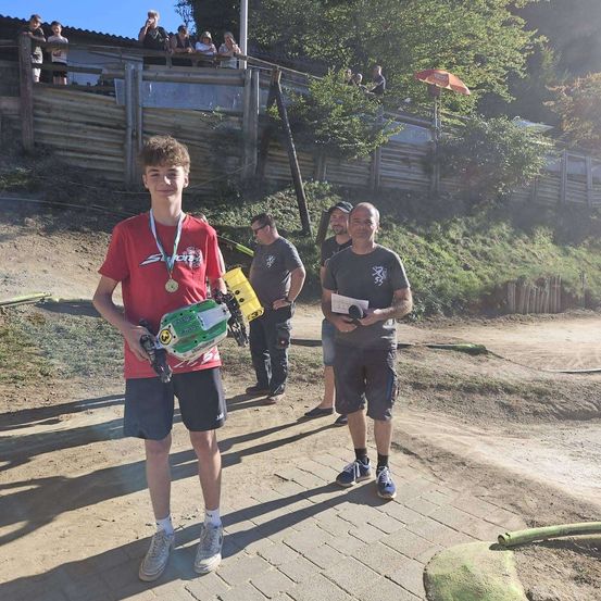 Ein junger Junge steht auf einem Lehmweg, hält ein ferngesteuertes Auto und eine Medaille und zwei Männer stehen hinter ihm, einer hält ein Mikrofon.