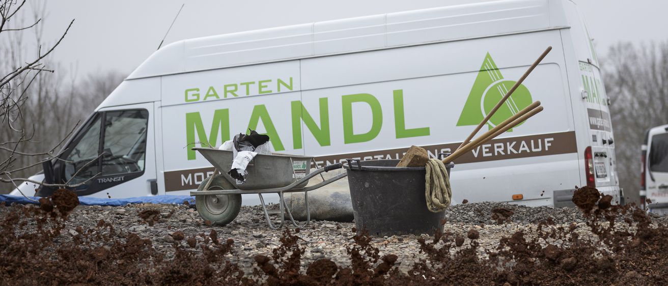 A white van with the text Garten Mandl in green stands next to a wheelbarrow and a bucket, both filled with soil.