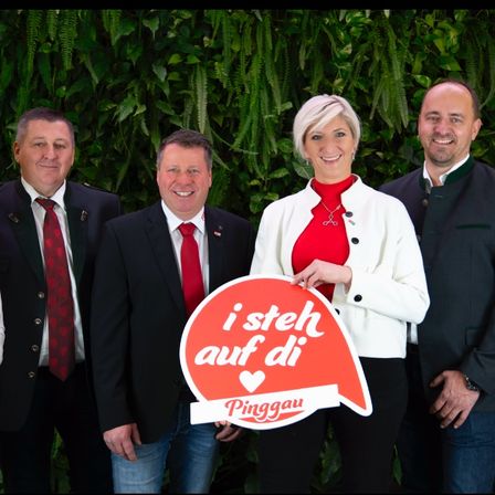 Fünf Personen stehen vor einem grünen Hintergrund. Eine Frau hält ein Schild mit der Aufschrift 'i steh auf di Pinggau'. Alle lächeln.