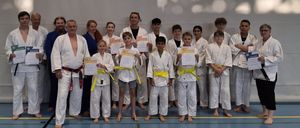 A group of martial arts students poses with certificates in hand, wearing white uniforms and belts, in a sports hall.