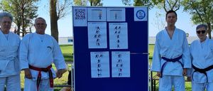 Five individuals in white martial arts uniforms stand in front of a blue board with exercise diagrams and a QR code. They are on a grassy field with trees in the background.