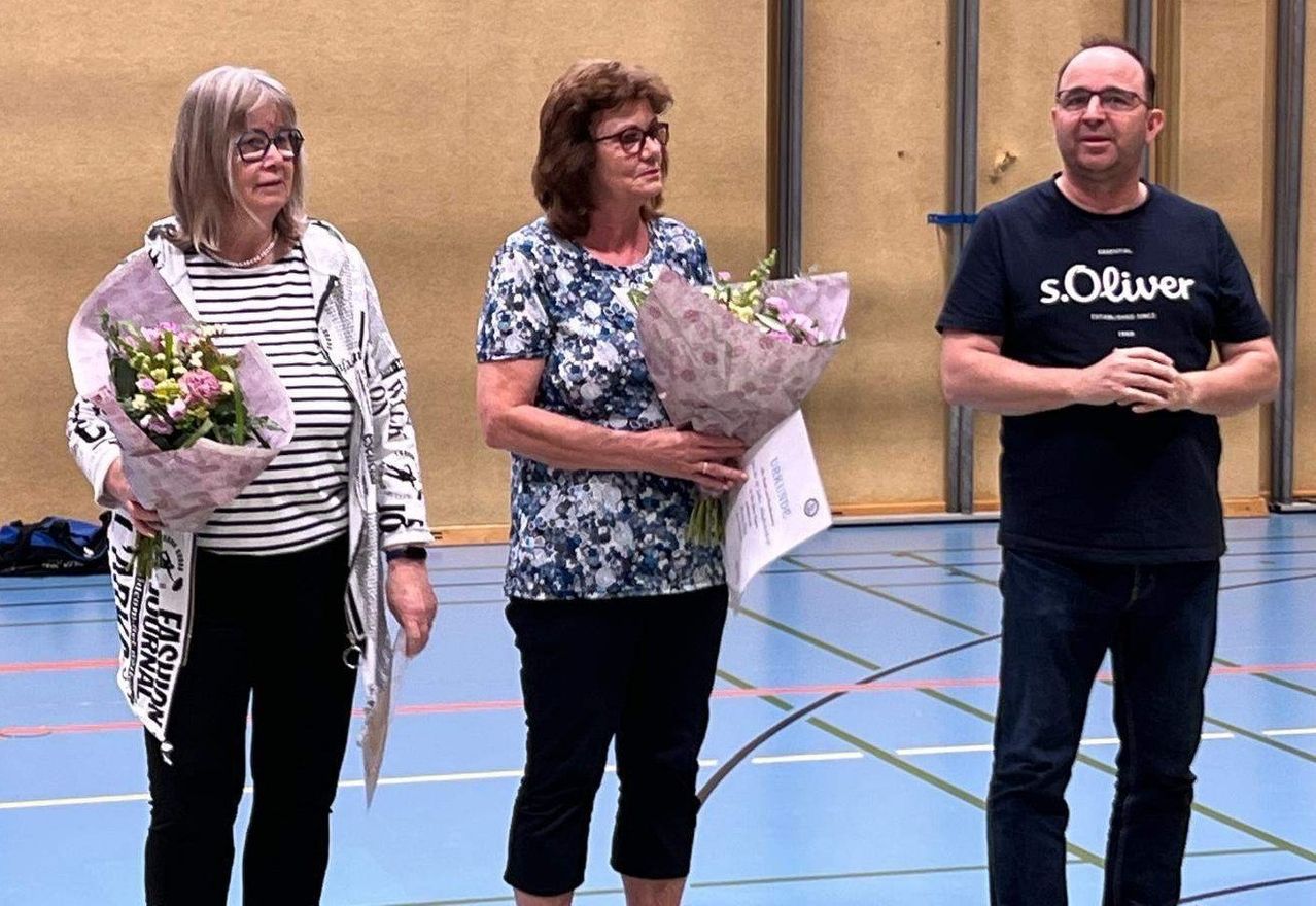 Drei Menschen stehen in einer Sporthalle und halten Blumen. Die Frau in der Mitte hält einen Blumenstrauß, während die anderen kleinere Sträuße halten. Die Frau rechts trägt eine Brille und einen Ring.