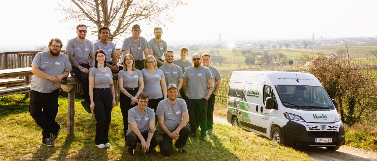 Eine Gruppe von Personen, möglicherweise ein Team, steht und sitzt auf einem Grasfeld und posiert für ein Gruppenfoto. Hinter ihnen steht ein Baum und ein Lieferwagen, der an der Seite geparkt ist. Sie tragen alle graue T-Shirts und Turnschuhe. Der Himmel ist klar und hell.