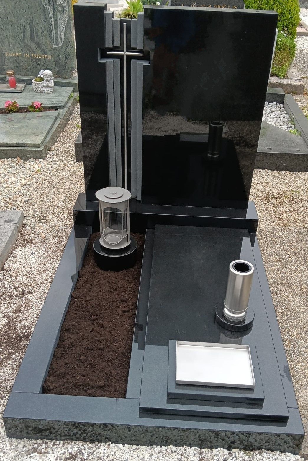 Ein schwarzer Grabstein mit zwei Metallröhren steht auf einem Friedhof. Auf beiden Seiten des Grabsteins befinden sich zwei Kerzenhalter. Eine kleine Silberplatte ist auf der rechten Seite platziert.