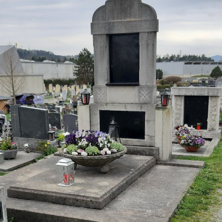 Ein Friedhof mit einem großen Steinmonument, geschmückt mit Blumen, umgeben von anderen Gräbern und einem Rasenbereich.