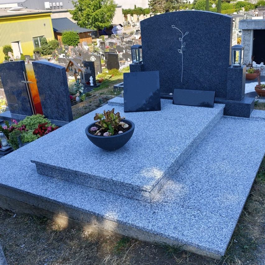 Ein grauer Granitgrabstein mit Blumendesign und einem Topf in einem Friedhof.