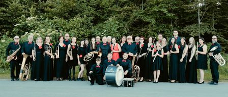 Eine große Gruppe von Musikern in schwarzer Kleidung mit roten Fliegen posiert für ein Foto im Freien, umgeben von Bäumen. Zu den Instrumenten gehören Trompeten, Posaunen, Tuben und Trommeln.