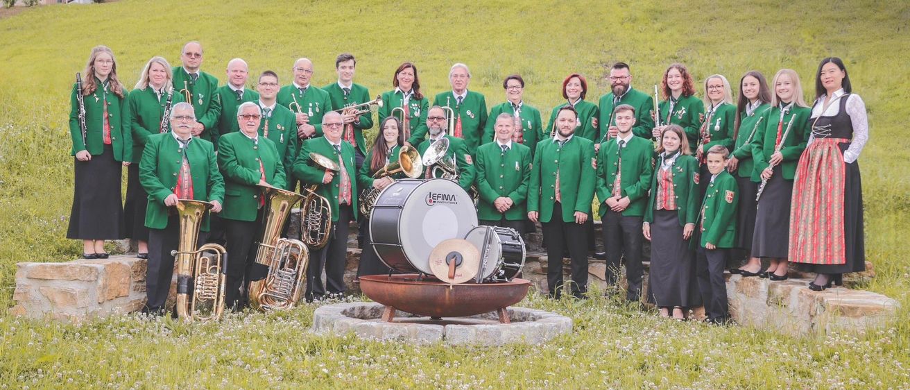 Eine Gruppe von Musikern in grünen Uniformen posiert mit ihren Instrumenten auf einem Rasenfeld. Im Vordergrund steht ein Schlagzeug in einer Steinschale.