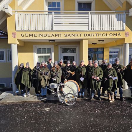 Eine Gruppe von Menschen in grünen Mänteln und Mützen steht vor einem Gebäude mit Balkon. Sie halten Musikinstrumente in den Händen, darunter Trompeten, Posaunen und eine Trommel. Das Gebäude trägt eine Beschilderung mit dem Namen Gemeindeamt Sankt Bartholomä.