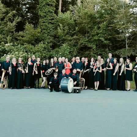 Eine Gruppe von Musikern in schwarzer und roter Kleidung posiert für ein Foto und hält verschiedene Instrumente, mit Bäumen im Hintergrund.