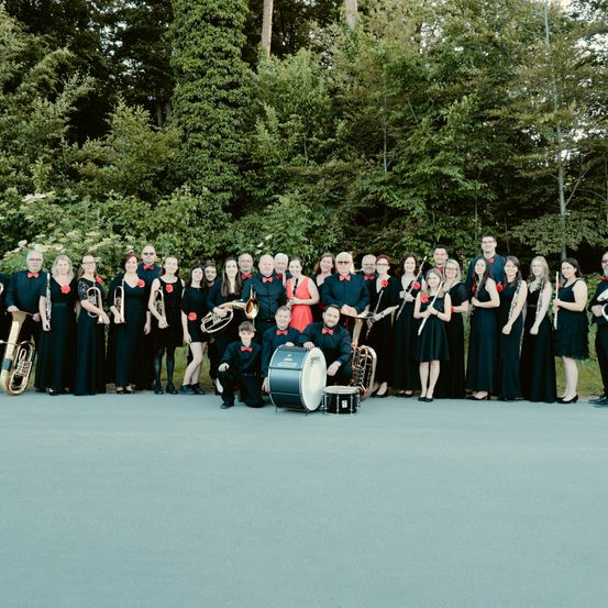 Eine Gruppe von Musikern in schwarzer und roter Kleidung posiert für ein Foto und hält verschiedene Instrumente, mit Bäumen im Hintergrund.