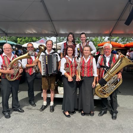 Eine Gruppe von Musikern in traditioneller deutscher Kleidung steht unter einem Zelt. Sie halten Blechblasinstrumente wie Tuba und Trompete. Zwei Frauen in der Mitte tragen Westen und schwarze Kleider. Dahinter sind verschwommene Menschen und Zelte zu sehen.