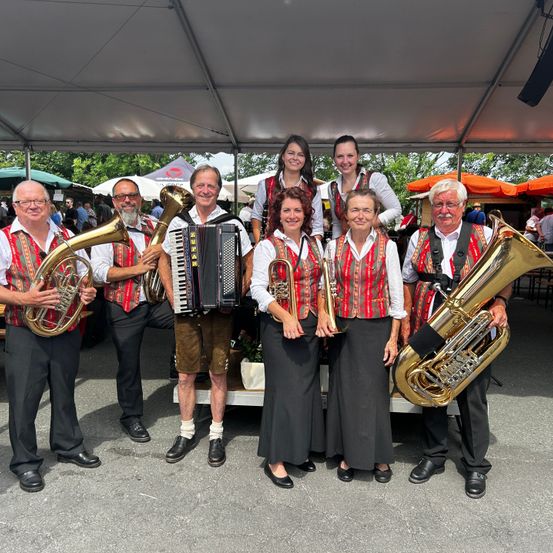 Eine Gruppe von Musikern in traditioneller deutscher Kleidung steht unter einem Zelt. Sie halten Blechblasinstrumente wie Tuba und Trompete. Zwei Frauen in der Mitte tragen Westen und schwarze Kleider. Dahinter sind verschwommene Menschen und Zelte zu sehen.