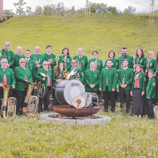 Eine große Gruppe von Musikern in grünen Uniformen steht auf einem grünen Hügel und posiert für ein Foto. Verschiedene Instrumente, einschließlich Trommeln, Tuben und Trompeten, sind vorhanden. Im Vordergrund befindet sich ein Steinsockel mit einem Feuerbecken.