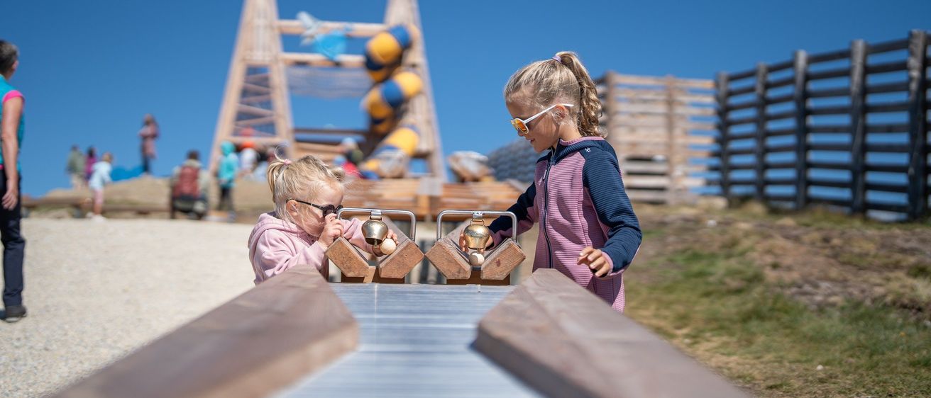 Zwei junge Mädchen spielen auf einer Rutsche mit Glocken in einem Outdoor-Spielplatz. Die Rutsche hat eine Metalloberfläche und ist von Holzstrukturen umgeben. Dahinter steht ein Holzzaun und eine Spiralrutsche.