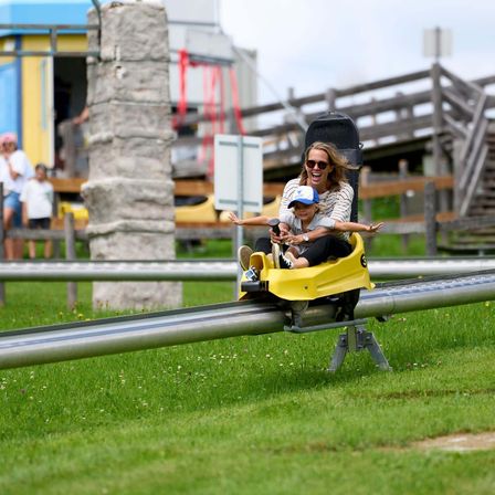 Eine Frau und ein Kind fahren eine gelbe Bobbahn auf einer Strecke. Beide haben Lächeln im Gesicht. Im Hintergrund stehen Menschen in der Nähe einer Holzstruktur.