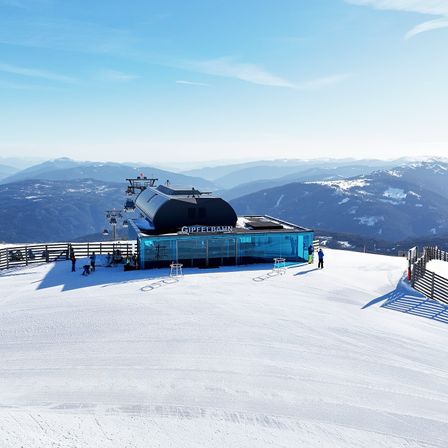 Eine Skiliftstation namens GIPFELBAHN befindet sich auf einem verschneiten Berg mit einem Zaun und Menschen um sie herum.
