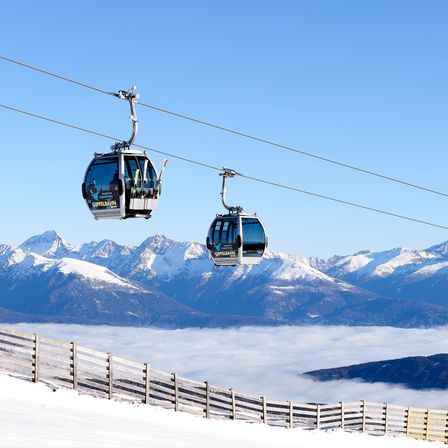 Zwei Seilbahnen fahren über einer verschneiten Landschaft mit Bergen und Nebel. Die Kabinen sind mit GIPFELBAHN beschriftet. Im Vordergrund befindet sich ein Holzzaun.
