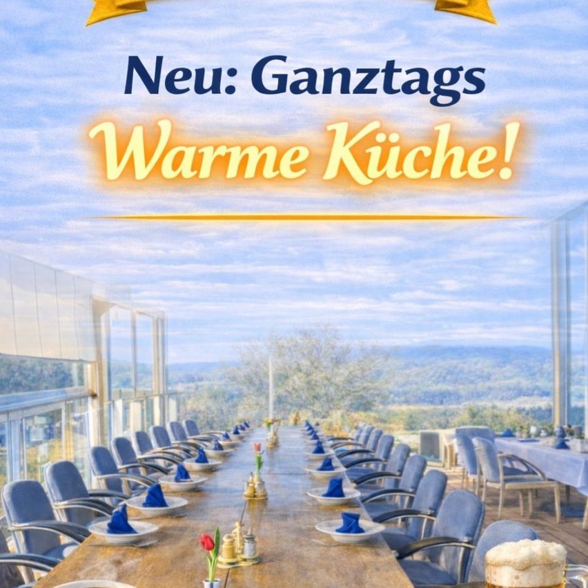 An outdoor restaurant with a long wooden table, neatly set with plates, blue napkins, and glasses. The view includes mountains and a blue sky. It says, 'New: Ganztags Warme Kuche!'.