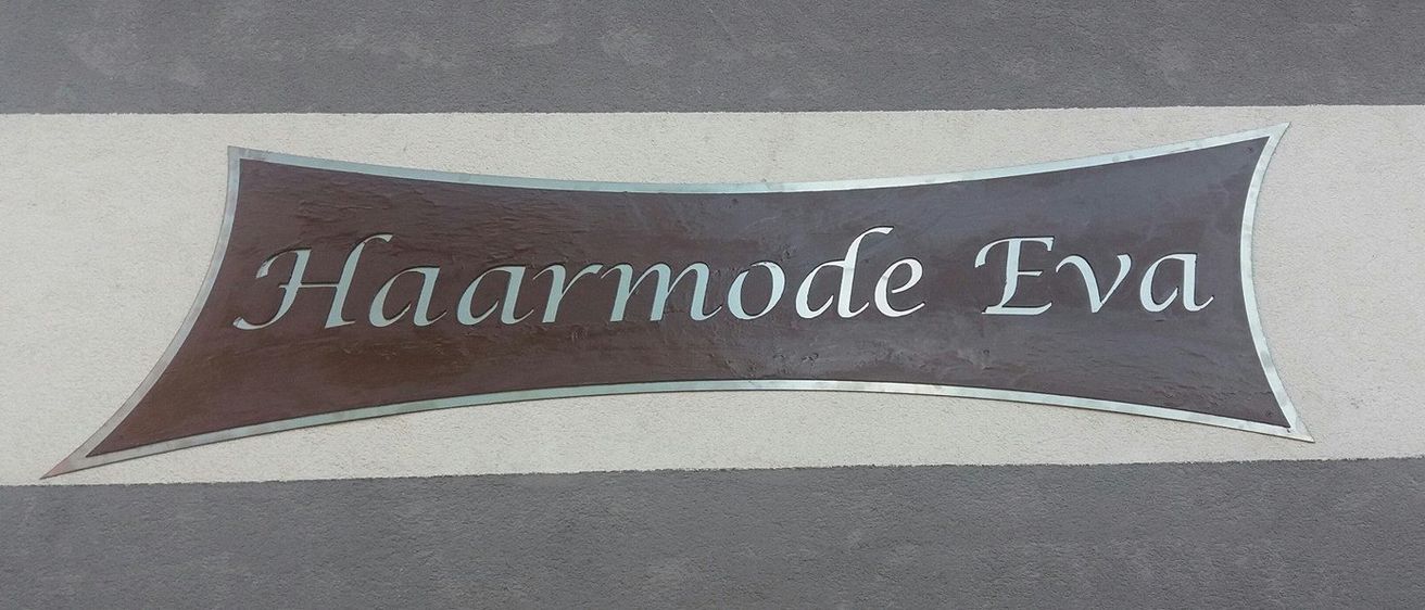 Ein braunes Schild mit weißen Buchstaben steht 'Haarmode E'. Das Schild ist an einer Wand mit grauen und weißen Streifen montiert.