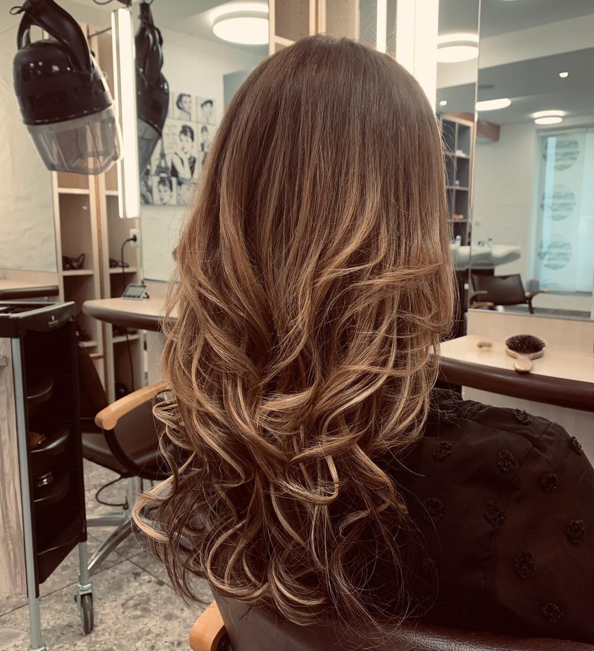 Eine Frau mit langen, welligen blonden Haaren sitzt in einem Salon, dem Kamerarücken zugewandt, mit einem Haartrockner an der Wand hinter ihr.