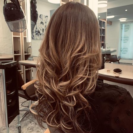 Eine Frau mit langen, welligen blonden Haaren sitzt in einem Salon, dem Kamerarücken zugewandt, mit einem Haartrockner an der Wand hinter ihr.