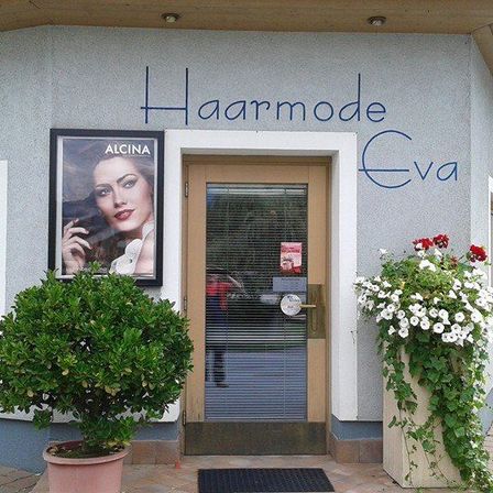 Das Äußere eines Gebäudes zeigt den Namen Haarmode Eva in Blau, mit einem Bilderrahmen, der das Porträt einer Frau zeigt, und Topfpflanzen auf beiden Seiten des Eingangs.
