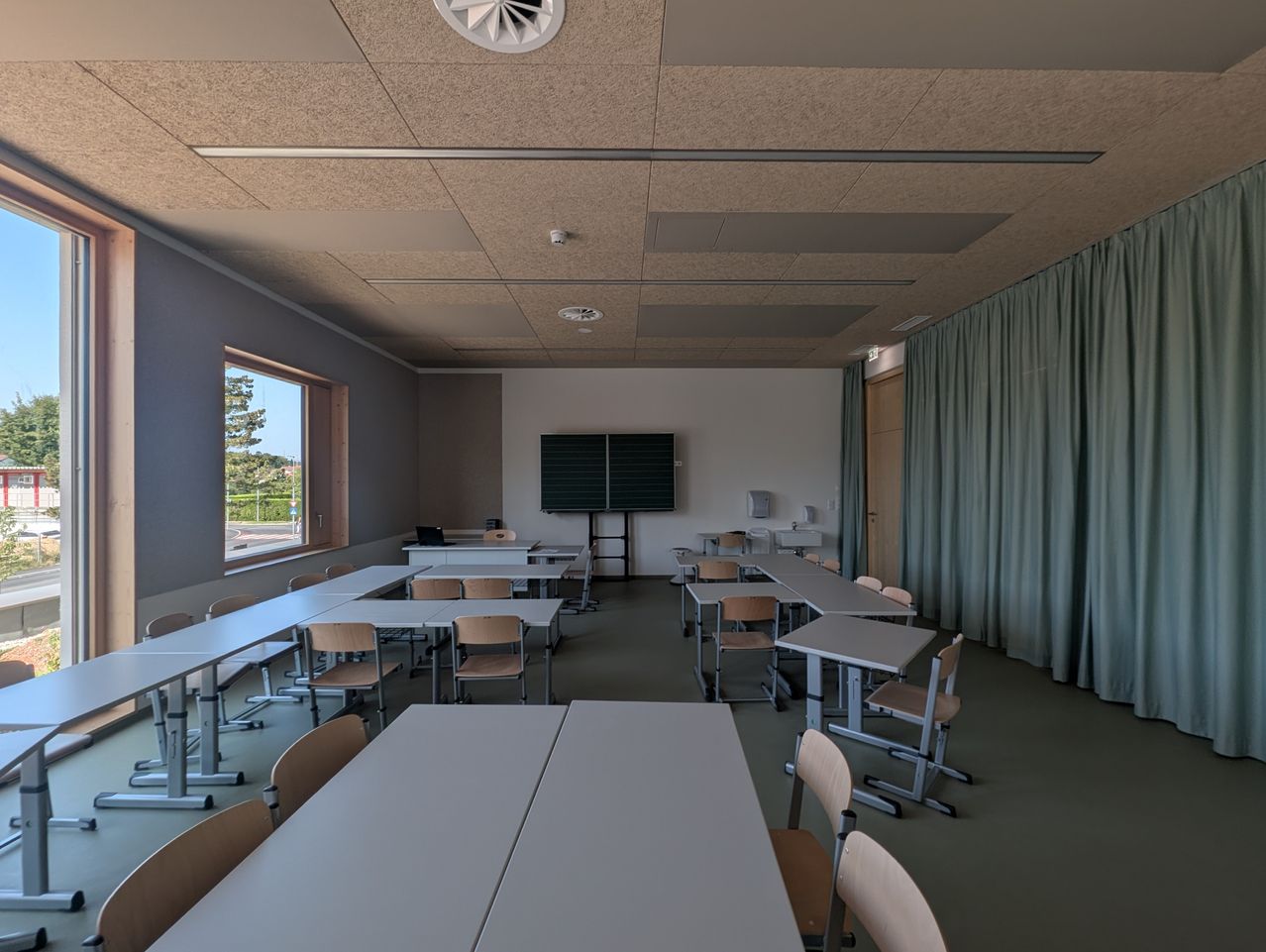 Ein leerer Klassenraum mit in Reihen angeordneten Tischen und Stühlen. Ein großes Fenster lässt natürliches Licht herein, und ein Fernseher ist an der Wand montiert. Die Decke hat ein Belüftungssystem.