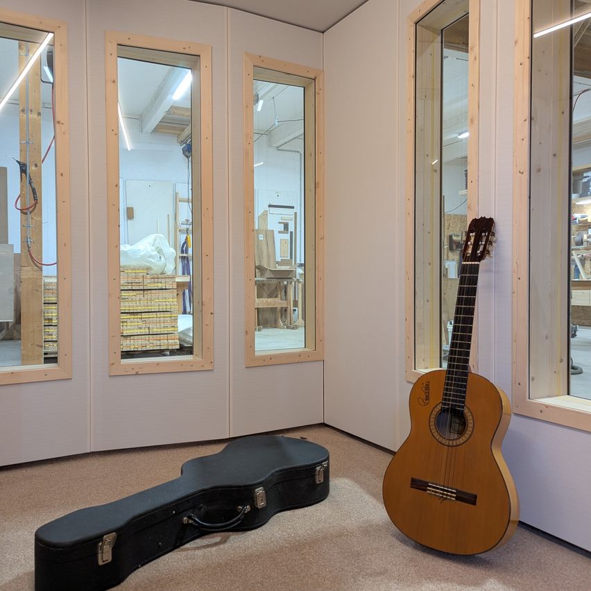 Eine Gitarre steht an einer Wand mit einem Holzrahmen, wobei ihre Hülle neben ihr auf dem Boden liegt. Der Raum hat große Glasfenster und wird von Lampen beleuchtet.