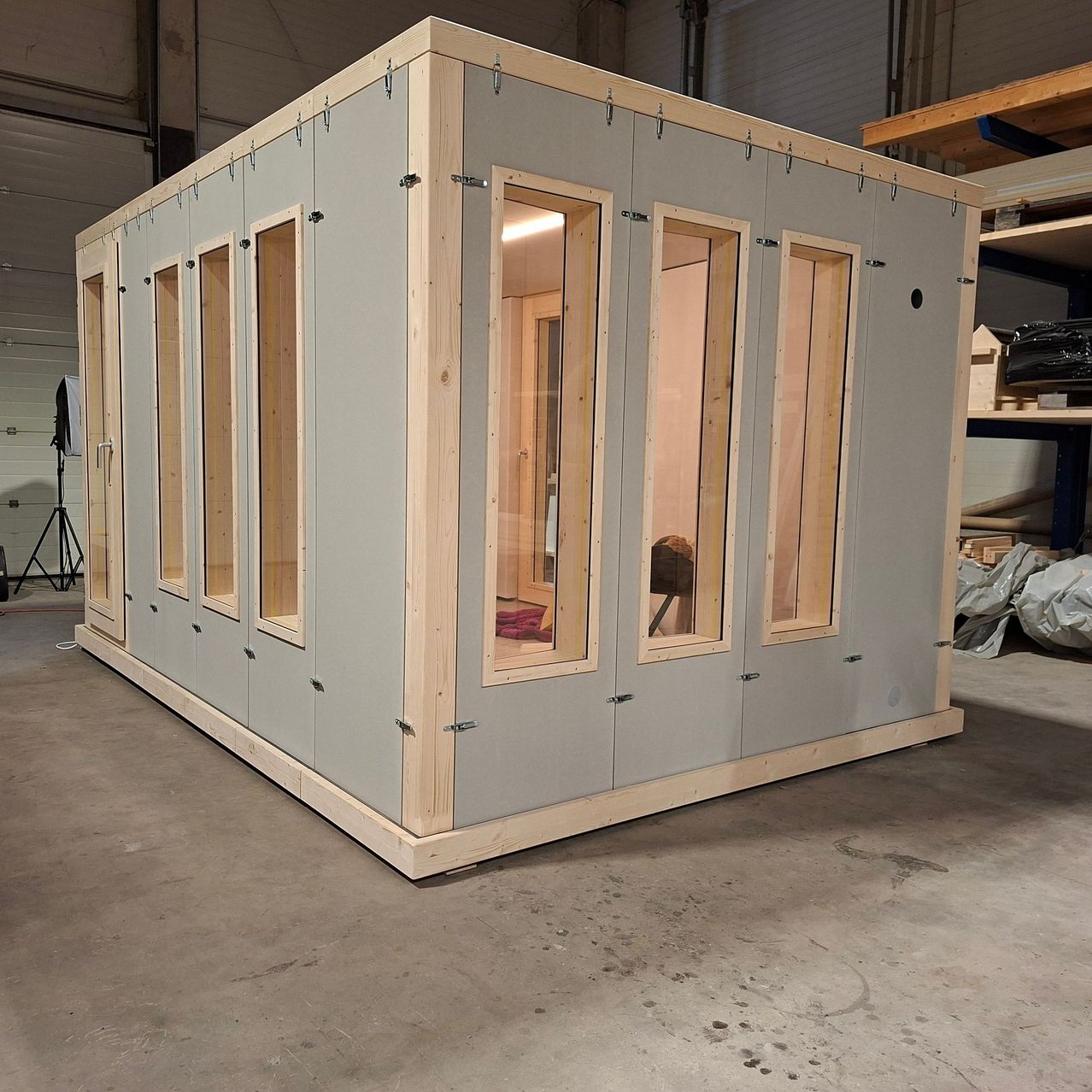 Ein unfertiger modularer Raum mit Holzrahmen und Glasfenstern befindet sich in einem Lagerhaus. Mehrere Fenster sind offen, und in der Nähe befindet sich ein Holzregal.