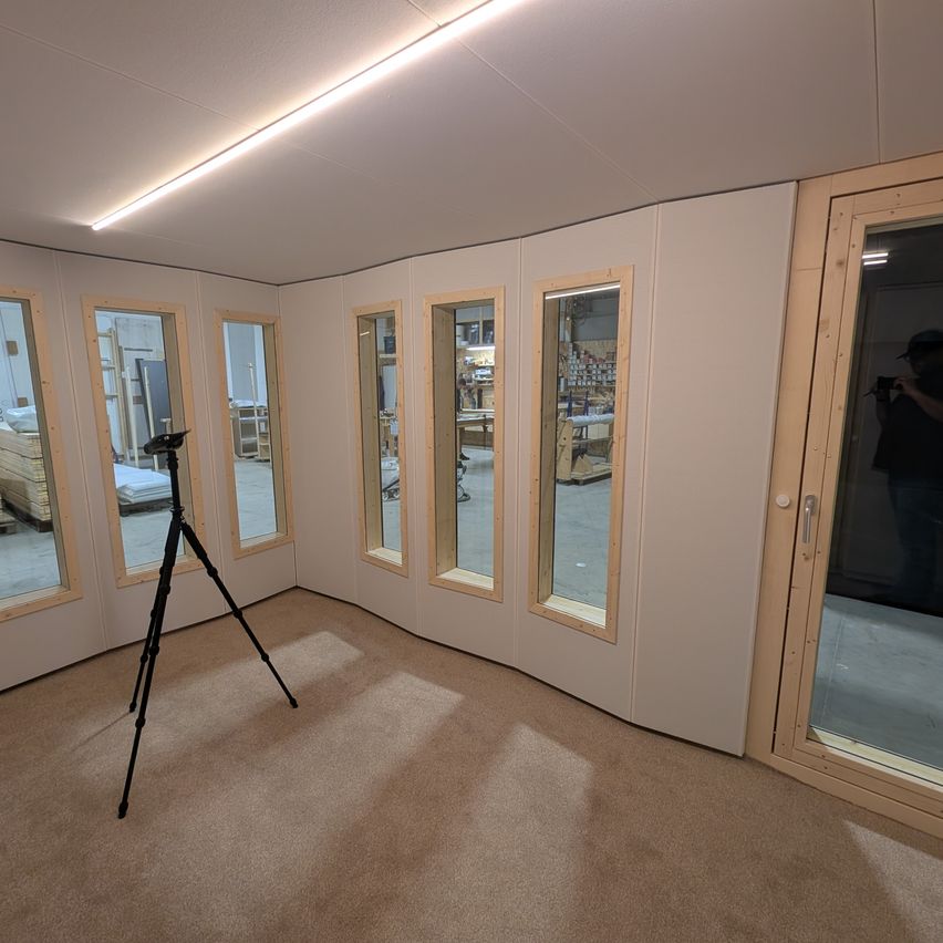 Ein leerer Raum mit weißen Wänden, Teppichboden und einem aufgestellten Stativ. Es gibt mehrere große Fenster mit Holzrahmen. Draußen steht eine Person und macht ein Foto.