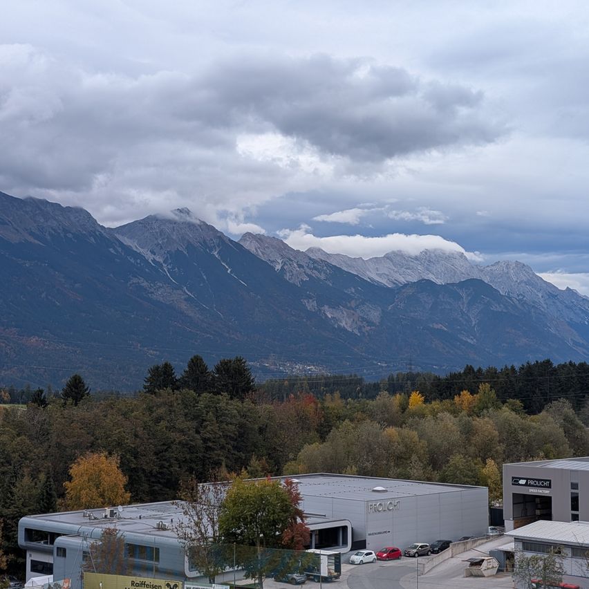 Eine Bergkette mit schneebedeckten Gipfeln unter einem bewölkten Himmel. Im Vordergrund sind mehrere Autos vor einem großen Industriegebäude geparkt.