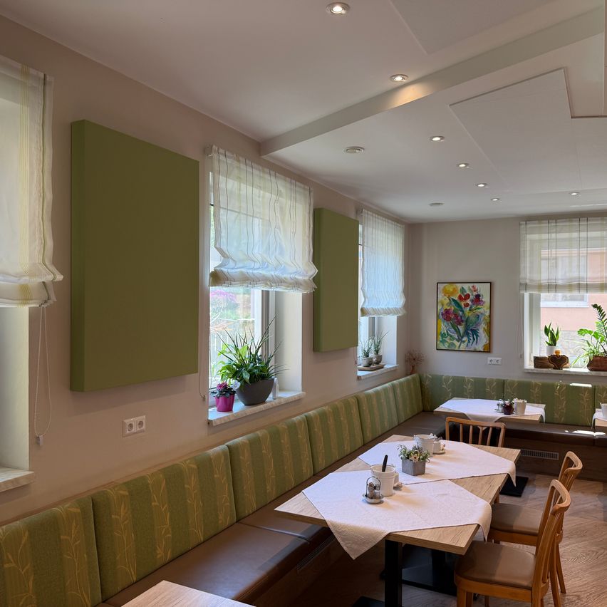 Ein gemütliches Restaurant mit grünen Wänden, weißen Vorhängen und einem Tisch, der mit weißen Tischdecken, Topfpflanzen und einem gerahmten Gemälde an der Wand gedeckt ist.