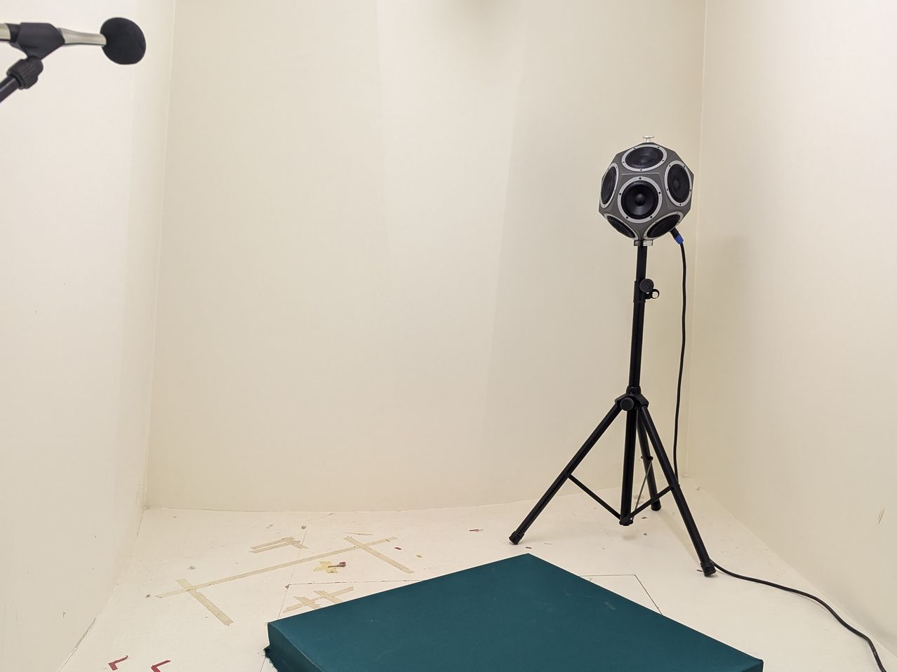 A spherical speaker is mounted on a tripod in a corner of a room. A green cushion is on the floor.
