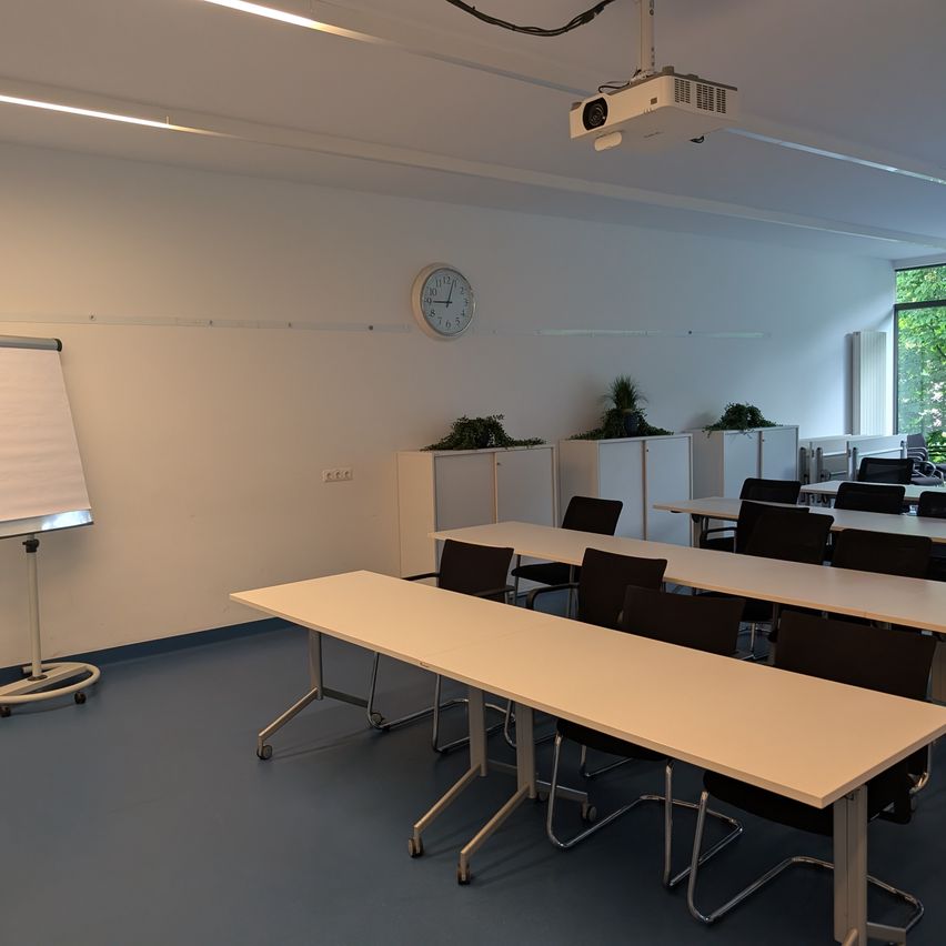 An empty classroom with white walls, a whiteboard, a projector, and several desks and chairs arranged in rows. The room is well-lit and has a clock on the wall.