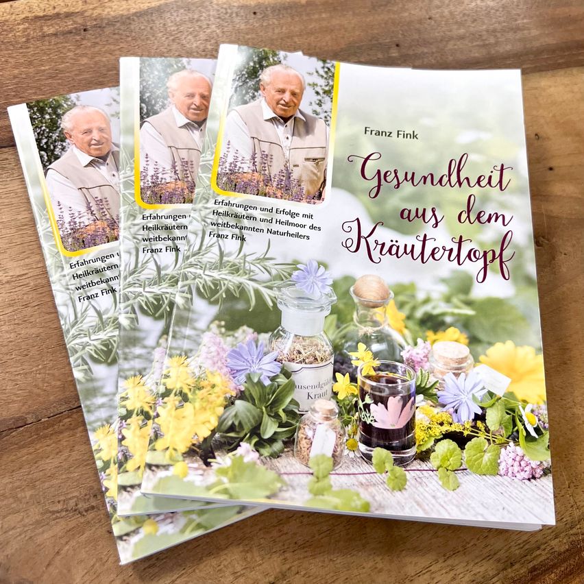 Drei Exemplare eines Buches mit dem Titel 'Gesundheit aus dem Krautertopf' liegen auf einer Holzoberfläche. Das Buch zeigt ein Bild eines Mannes in einem weißen Weste und einem Stapel kleiner Gläser.