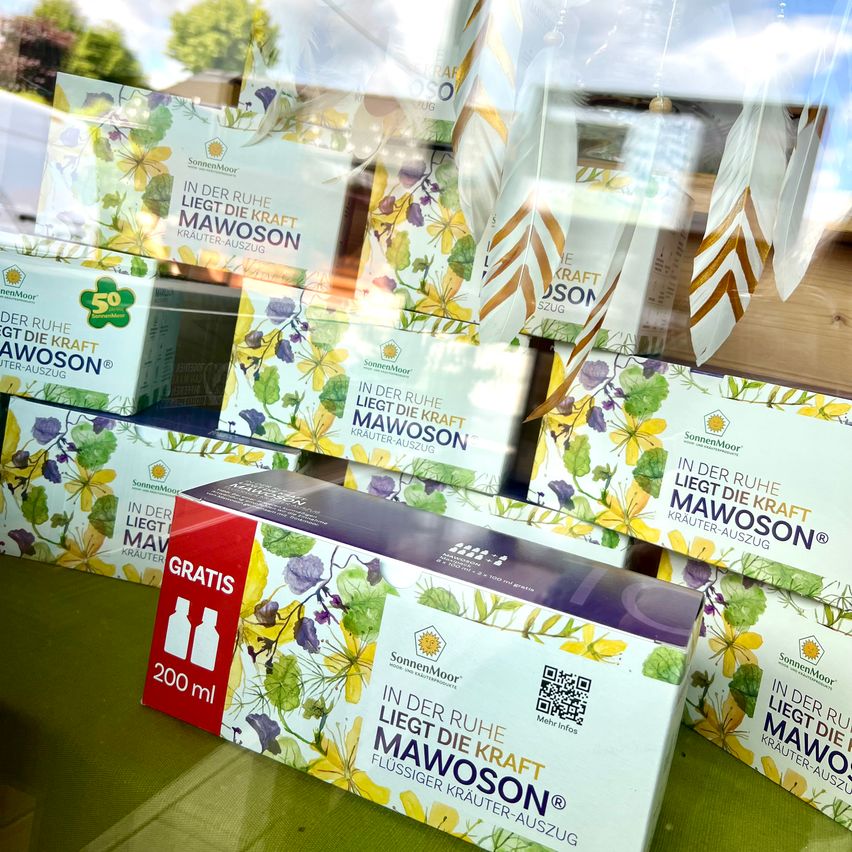 Eine Ausstellung von Schachteln mit Kräuterextrakten, beschriftet mit 'In der Ruhe liegt die Kraft Mawoson', mit einer roten Schachtel, die 'GRATIS 200 ml' anzeigt, vorne, auf einer grünen Oberfläche angeordnet.