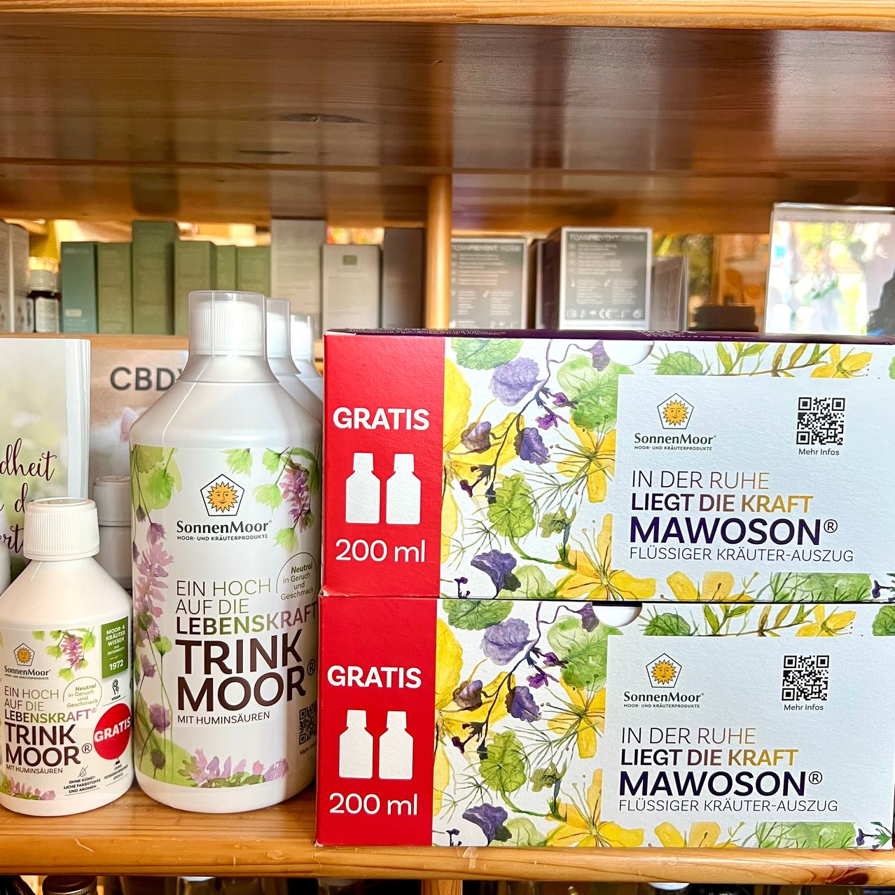 Auf einem Holzregal sind mehrere Flaschen und Schachteln von SonnenMoor Produkten ausgestellt. Es gibt zwei weiße Flaschen und zwei Schachteln, eine mit der Aufschrift 'GRATIS' und einem QR-Code. Die Schachteln haben leuchtende Blumendesigns und sind mit 'SonnenMoor' und 'MAWOSON' beschriftet.