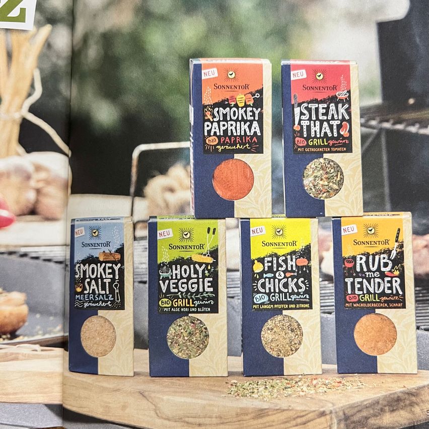 Ein Holzschneidebrett hält mehrere Schachteln biologischer Gewürze von Sonnentor. Dazu gehören Smokey Paprika, Steak That, Smokey Salt, Holy Veggie, Fish & Chicks und Rub Me Tender.