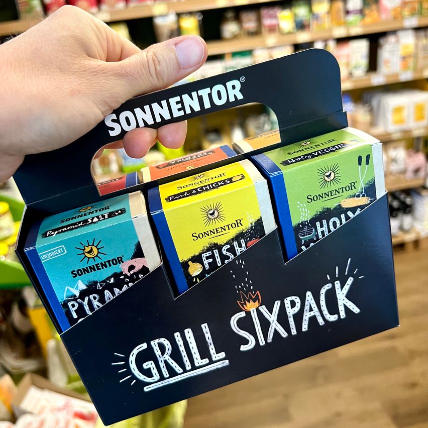 Eine Person hält eine schwarze Box mit der Aufschrift 'Sonnentor Grill Sixpack', die vier Gewürzpackungen enthält: Pyramid Salt, Fish & Chicks, Holy Veggie und Holy Smoke.