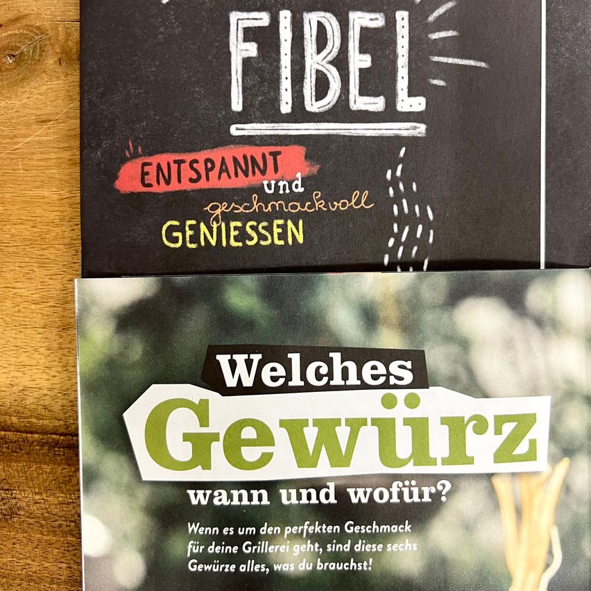 Zwei Kochbücher auf einer Holzoberfläche. Das obere hat 'Fibel' in weißen und roten Buchstaben mit den Worten 'Entspannt und Geschmackvoll Geniessen'. Darunter ein zweites Buch mit dem Titel 'Welches Gewürz wann und wofür?' mit einem verschwommenen grünen Hintergrund.