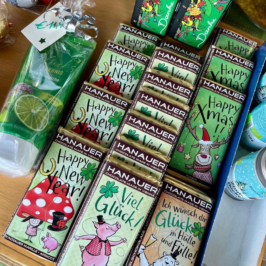 Auf einem Holztisch steht eine Schachtel mit Schokoladen mit festlichen Grüßen, darunter Happy New Year und Viel Gluck. Die Schachtel ist blau mit einem grünen Deckel und enthält eine kleine Packung Limette und eine Karte mit einem Geschenkanhänger.